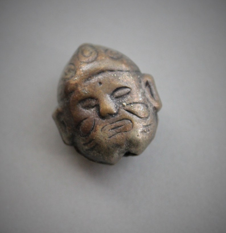 May include: A small, detailed, bronze-colored figurine of a man's face with a patterned hat. The figurine has a weathered look and appears to be made of metal.