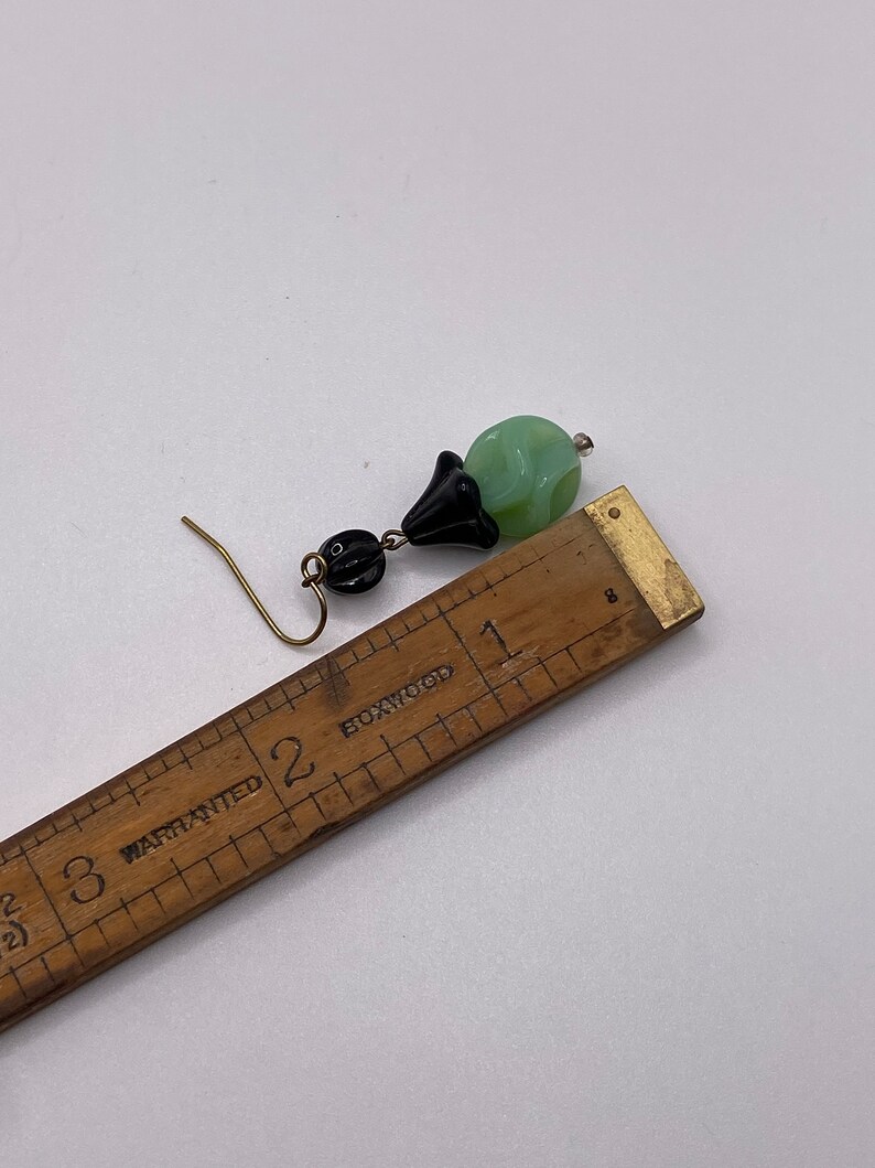 May include: A pair of dangle earrings with a green and black glass bead and a black metal flower charm. The earrings are approximately 2 inches long.