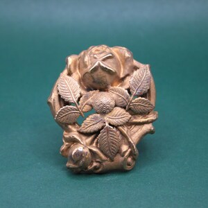 May include: A gold-colored, vintage-style brooch featuring a detailed rose design. The brooch has a central rose bud surrounded by detailed leaves. The metal has a slightly aged appearance, suggesting it is an antique or vintage piece.
