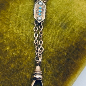 May include: A silver pocket watch chain with a decorative fob featuring turquoise and pearl accents. The chain has a swivel clasp.