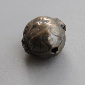May include: A small, round, bronze bell with intricate carvings. The bell has a small opening at the top for hanging.