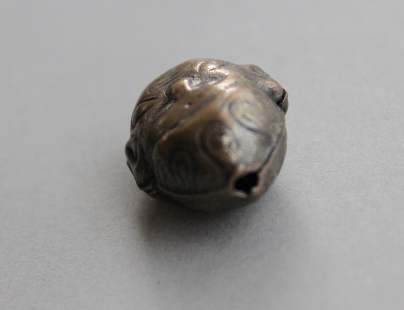 May include: A small, round, bronze bell with intricate carvings. The bell has a small opening at the top for hanging.