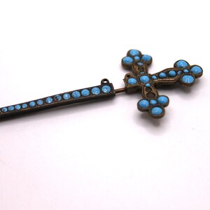 May include: An antique cross brooch with a pin. The brooch has a bronze-coloured metal frame and is decorated with turquoise-coloured stones. The cross design has rounded edges and is a religious accessory.