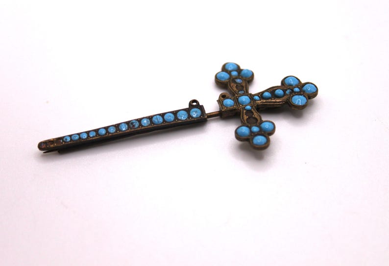 May include: An antique cross brooch with a pin. The brooch has a bronze-coloured metal frame and is decorated with turquoise-coloured stones. The cross design has rounded edges and is a religious accessory.