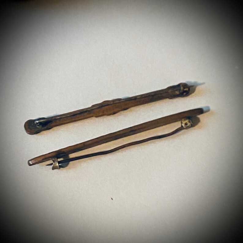 May include: Two gold-toned metal brooches with a simple design. One brooch is straight and the other is slightly curved.