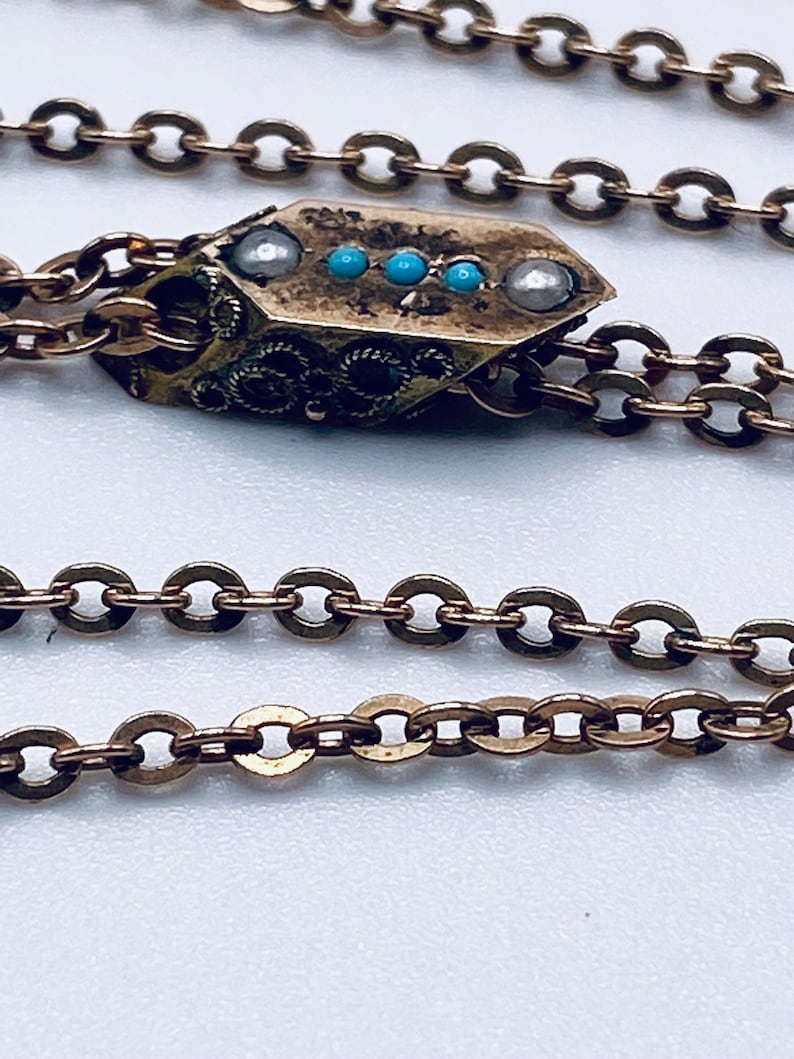 May include: A gold chain necklace with a decorative pendant. The pendant is rectangular in shape with a textured surface and features small blue and white stones.