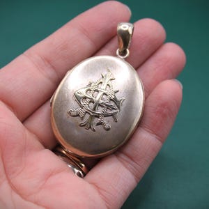 May include: Oval gold locket with a decorative monogram. The locket has a loop at the top for a chain and a hinged opening. The monogram is in a contrasting gold colour and is centered on the front of the locket.