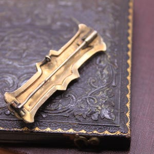 May include: A vintage gold-toned brooch with an ornate design, featuring a pin on the back. The brooch is set against a dark, textured surface, possibly leather, with a decorative border. The overall aesthetic suggests a classic, antique style.