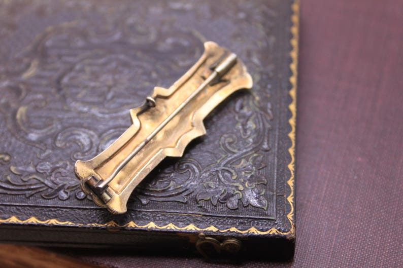 May include: A vintage gold-toned brooch with an ornate design, featuring a pin on the back. The brooch is set against a dark, textured surface, possibly leather, with a decorative border. The overall aesthetic suggests a classic, antique style.