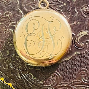 May include: A round gold-toned metal locket featuring an ornate, engraved monogram with the intertwined letters E and F on the front surface. The locket includes a small circular bail at the top for attachment to a chain or cord.