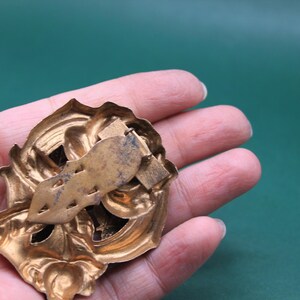 May include: Ornate gold-colored metal brooch with a decorative floral design. The brooch has a detailed, textured surface and a pin clasp on the back. The brooch is held in a hand against a green background.