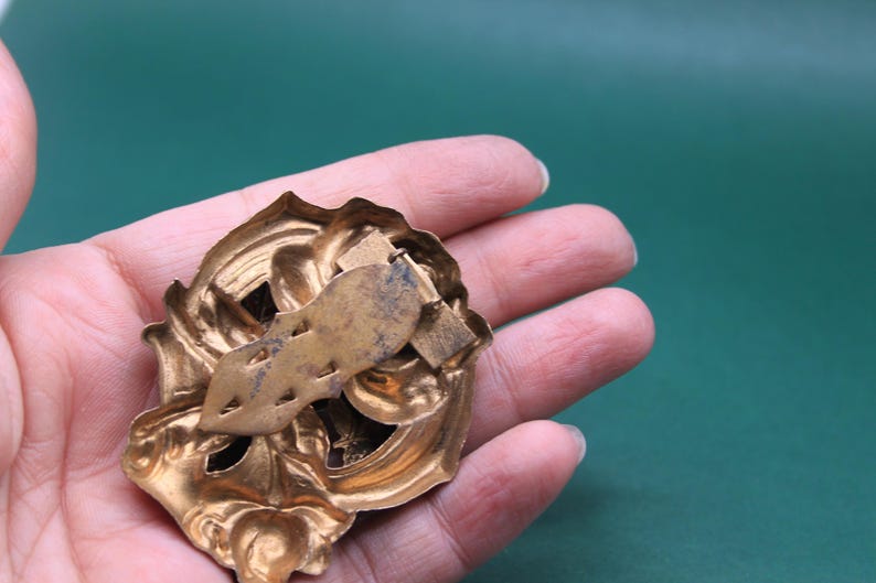 May include: Ornate gold-colored metal brooch with a decorative floral design. The brooch has a detailed, textured surface and a pin clasp on the back. The brooch is held in a hand against a green background.