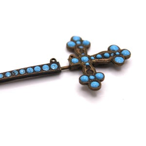 May include: An antique bronze-coloured cross pendant with turquoise-coloured stones. The cross features a detailed design with rounded stones and a matching bar with similar stones. The pendant is set against a white background.