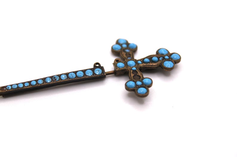 May include: An antique bronze-coloured cross pendant with turquoise-coloured stones. The cross features a detailed design with rounded stones and a matching bar with similar stones. The pendant is set against a white background.