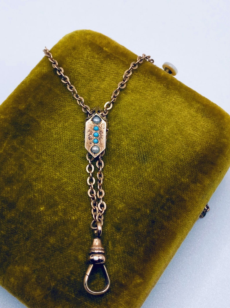 May include: A gold chain watch fob with a decorative pendant featuring turquoise and pearl accents. The fob is on a green velvet background.