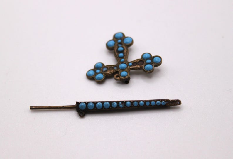 May include: An antique brooch and pin set. The cross-shaped brooch and the pin are both bronze-toned and decorated with turquoise-coloured stones. The cross has rounded edges and the pin is a straight bar with a series of turquoise stones.