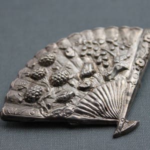 May include: A silver-toned fan-shaped brooch with intricate floral and leaf designs. The brooch features a raised, textured surface and a detailed fan design.