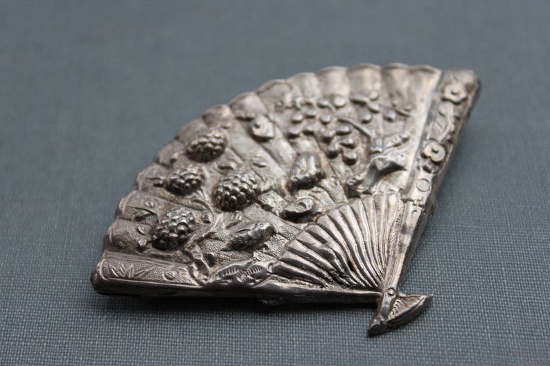 May include: A silver-toned fan-shaped brooch with intricate floral and leaf designs. The brooch features a raised, textured surface and a detailed fan design.