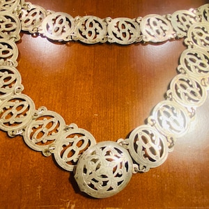 May include: A silver-toned metal belt with an ornate, openwork design. The belt features a series of connected oval and circular links, each with a decorative pattern. The central clasp is an oval shape with a floral design. The belt is displayed on a wooden surface.