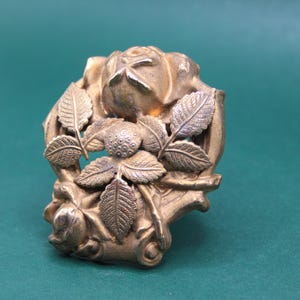 May include: A detailed, antique-style gold-colored metal brooch. The brooch features a central rose with surrounding leaves and floral accents. The design is intricate and ornate, with a vintage aesthetic.