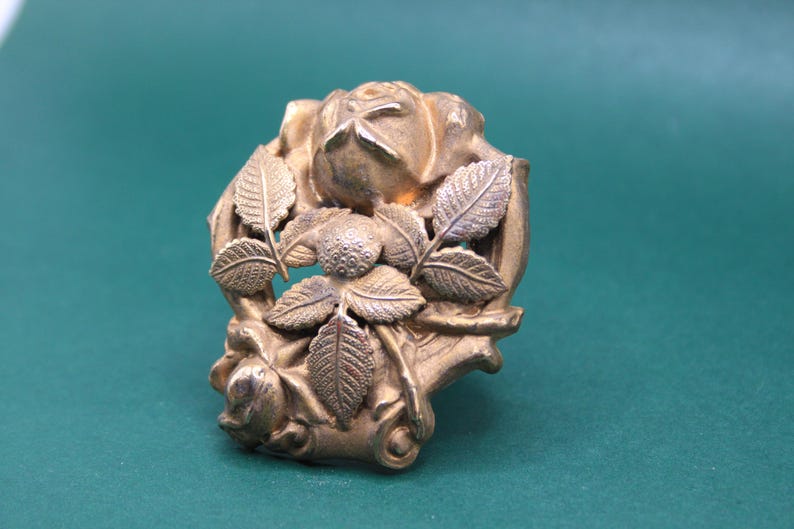 May include: A detailed, antique-style gold-colored metal brooch. The brooch features a central rose with surrounding leaves and floral accents. The design is intricate and ornate, with a vintage aesthetic.