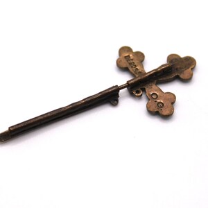 May include: A vintage bronze cross-shaped pin. The cross features four rounded terminals and a long, thin pin. The word "DEROSA" is inscribed on the cross. The pin is set against a white backdrop.
