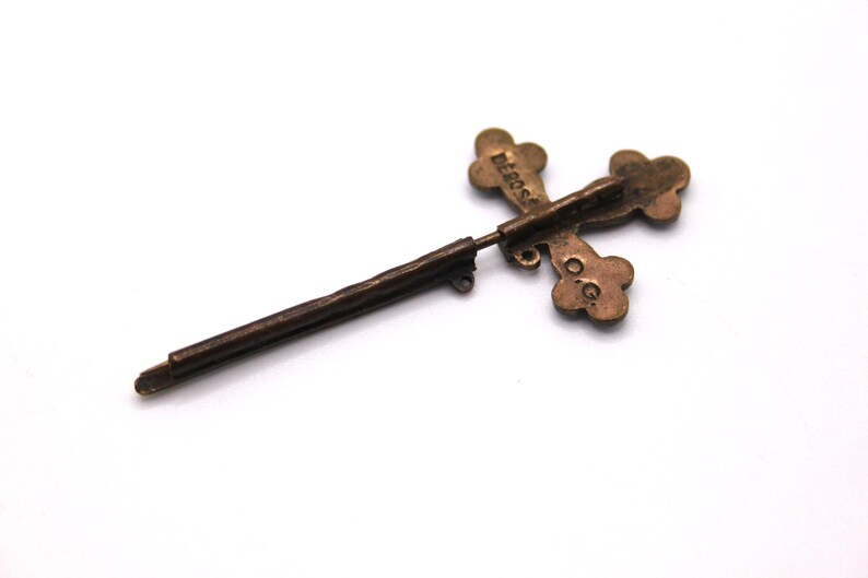 May include: A vintage bronze cross-shaped pin. The cross features four rounded terminals and a long, thin pin. The word "DEROSA" is inscribed on the cross. The pin is set against a white backdrop.