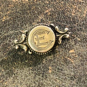 May include: Antique silver-toned brooch with an engraved letter "E" in a circular frame. The brooch features ornate scrollwork on either side of the central design, creating a vintage aesthetic. The brooch is likely made of a metal alloy.