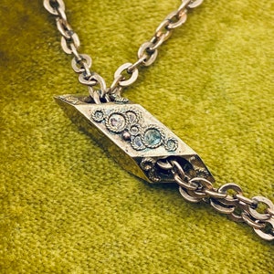 May include: A silver chain necklace with a decorative pendant. The pendant is shaped like a diamond and has a detailed design with small stones.