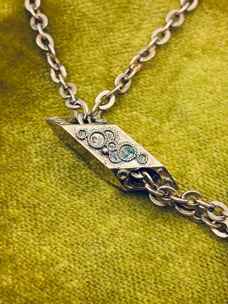 May include: A silver chain necklace with a decorative pendant. The pendant is shaped like a diamond and has a detailed design with small stones.