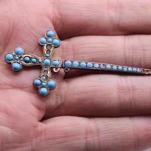 May include: A vintage cross brooch with a gold-toned metal frame and light blue turquoise stones. The cross features a detailed design with rounded edges and a pin closure. The brooch is a religious accessory, likely for clothing or a bag.