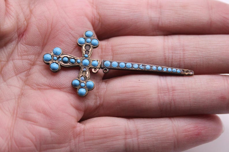 May include: A vintage cross brooch with a gold-toned metal frame and light blue turquoise stones. The cross features a detailed design with rounded edges and a pin closure. The brooch is a religious accessory, likely for clothing or a bag.