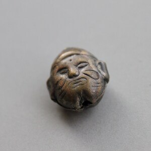 May include: A silver-colored bead with a detailed face carved into it. The face has a serious expression with a furrowed brow and a closed mouth.