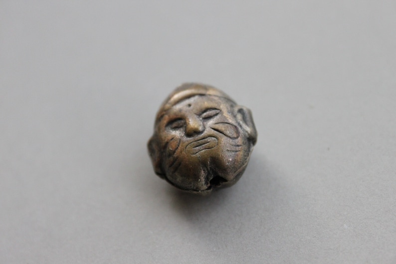 May include: A silver-colored bead with a detailed face carved into it. The face has a serious expression with a furrowed brow and a closed mouth.