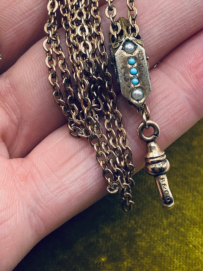 May include: A gold chain with a decorative pendant featuring turquoise and pearl accents. The chain has a fob with the inscription "B & CO."