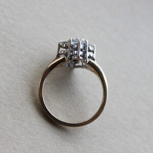 May include: A gold band ring with a large, clear, multi-faceted stone set in the center. The stone is surrounded by smaller, clear stones.
