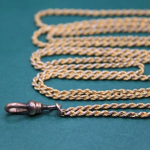 Puede incluir: Collar de cadena de cuerda dorada con cierre decorativo. La cadena está dispuesta en bucles sobre un fondo verde azulado. El cierre es una pequeña pieza de metal detallada. Joyería.
