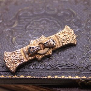 May include: An ornate gold-toned brooch with intricate filigree and a central clasp with black and gold accents. The brooch is set against a dark, textured surface with embossed floral patterns, suggesting a vintage or antique style.