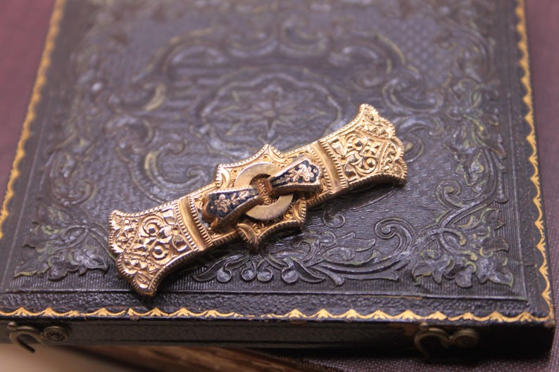May include: An ornate gold-toned brooch with intricate filigree and a central clasp with black and gold accents. The brooch is set against a dark, textured surface with embossed floral patterns, suggesting a vintage or antique style.