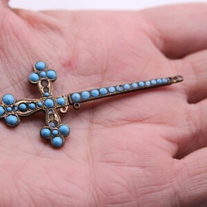 May include: A vintage cross-shaped brooch with a bronze-coloured metal frame and turquoise-coloured stone accents. The cross has a long, slender pin extending from one side, held in a person's hand.