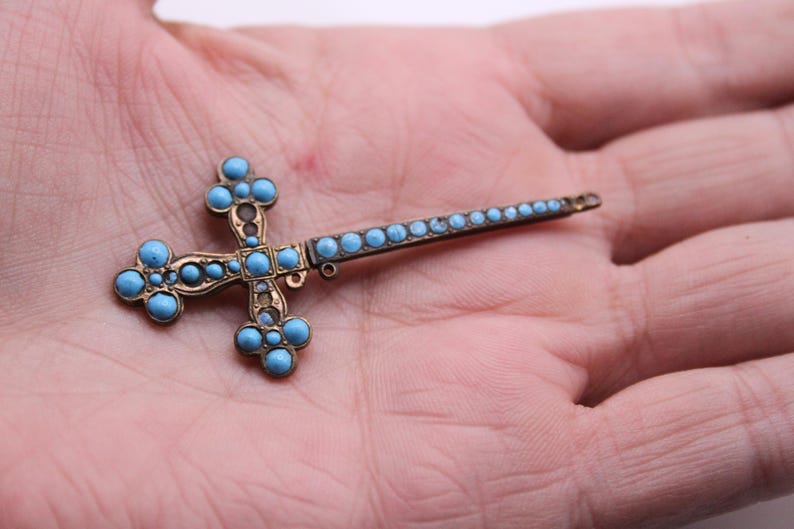 May include: A vintage cross-shaped brooch with a bronze-coloured metal frame and turquoise-coloured stone accents. The cross has a long, slender pin extending from one side, held in a person's hand.