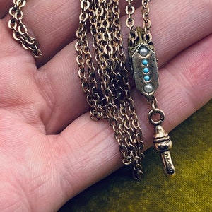 May include: A gold chain necklace with a pendant featuring a geometric design with turquoise and pearl accents. The pendant is marked with the letters "B & CO".