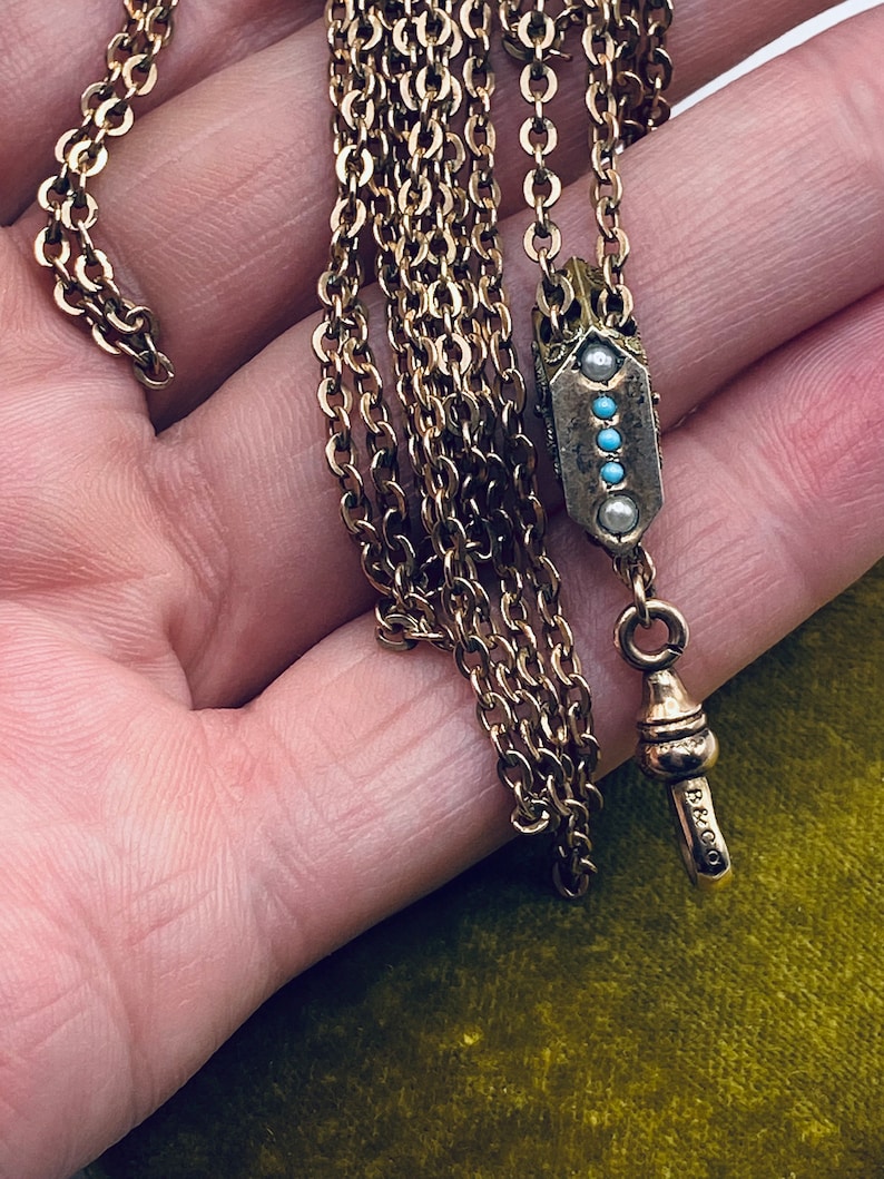 May include: A gold chain necklace with a pendant featuring a geometric design with turquoise and pearl accents. The pendant is marked with the letters "B & CO".