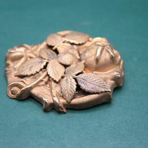 May include: A detailed, antique-style bronze-colored metal decorative piece. The design features leaves, berries, and a floral motif. The piece has a slightly irregular shape and is likely a decorative element.