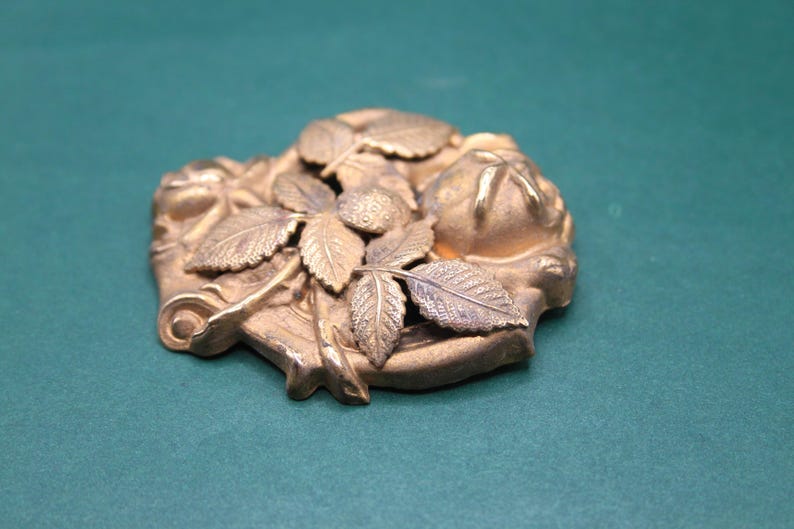 May include: A detailed, antique-style bronze-colored metal decorative piece. The design features leaves, berries, and a floral motif. The piece has a slightly irregular shape and is likely a decorative element.