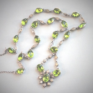 May include: A silver necklace featuring oval, light green gemstones and small, clear accent stones. The necklace has a delicate chain and a floral-shaped pendant at the bottom. The gemstones are evenly spaced along the chain.