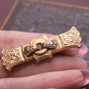 May include: A gold-toned antique brooch with intricate floral and scrollwork details. The brooch features a central bow design with black enamel accents and a decorative clasp. The ends of the brooch have ornate crown-like embellishments.