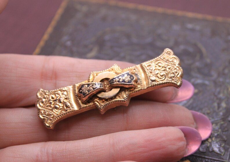 May include: A gold-toned antique brooch with intricate floral and scrollwork details. The brooch features a central bow design with black enamel accents and a decorative clasp. The ends of the brooch have ornate crown-like embellishments.