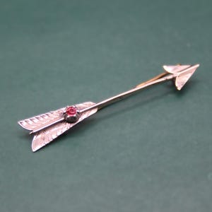 May include: A silver-toned arrow-shaped brooch with a red gemstone at the center. The arrow features detailed feathering and a pointed arrowhead. The brooch is set against a dark green background, highlighting its metallic sheen.