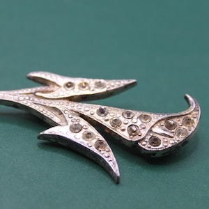 May include: A silver-toned brooch with a floral design. The brooch features three stylized, curved elements, each adorned with clear, round rhinestones. The brooch is set against a dark green background, highlighting its intricate details and vintage aesthetic.